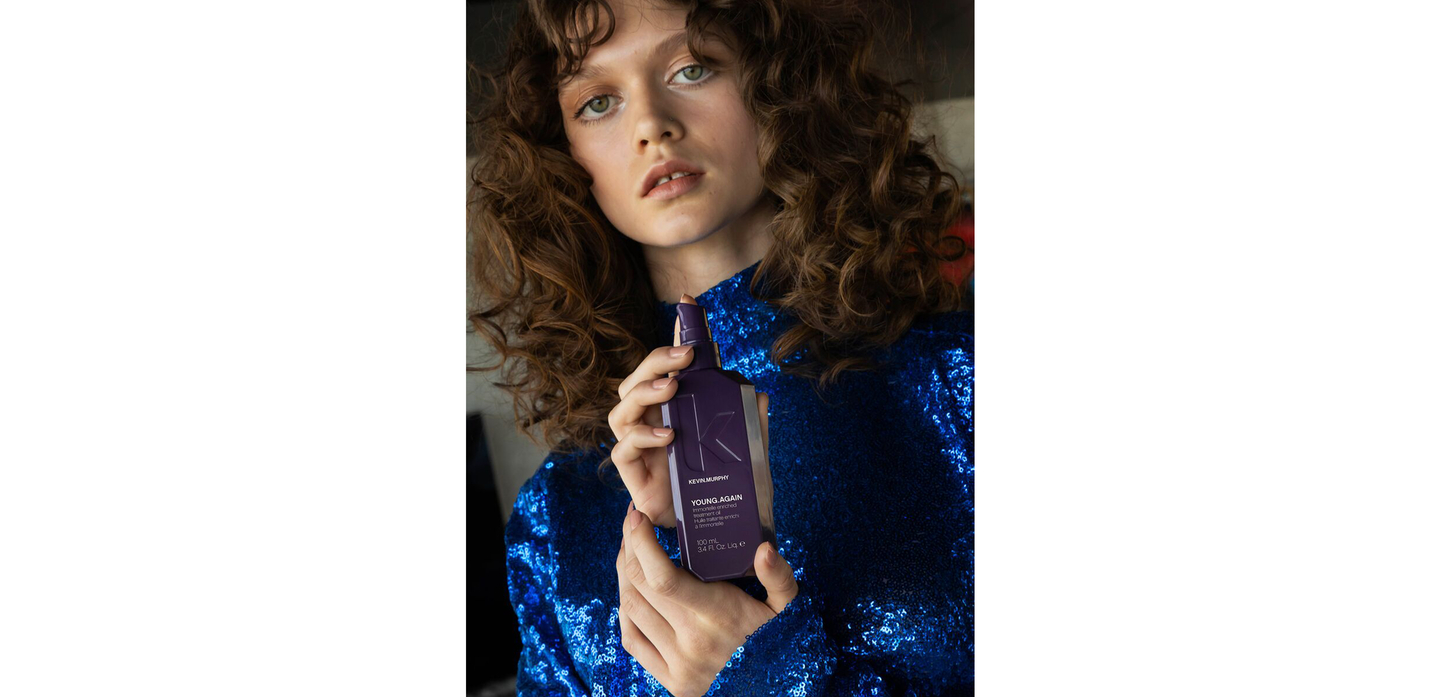 Woman with curly hair holding YOUNG.AGAIN treatment oil in blue sequin dress